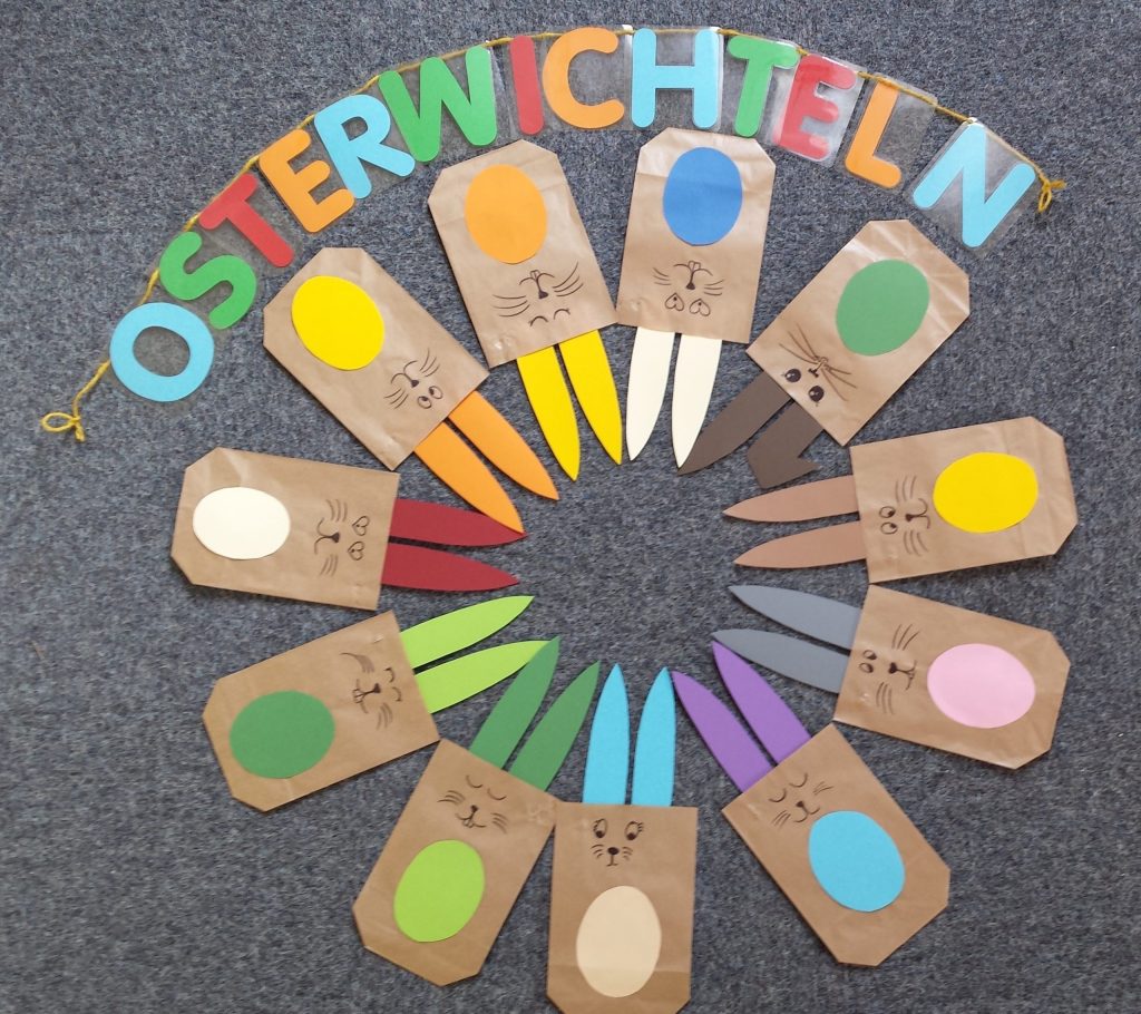 Gebastelte bunte Papiertüten Osterhasten liegen im Kreis. Oben drüber liegt im Halbkreis eine Buchstaben-Girlande. Sie schreibt OSTERWICHTELN