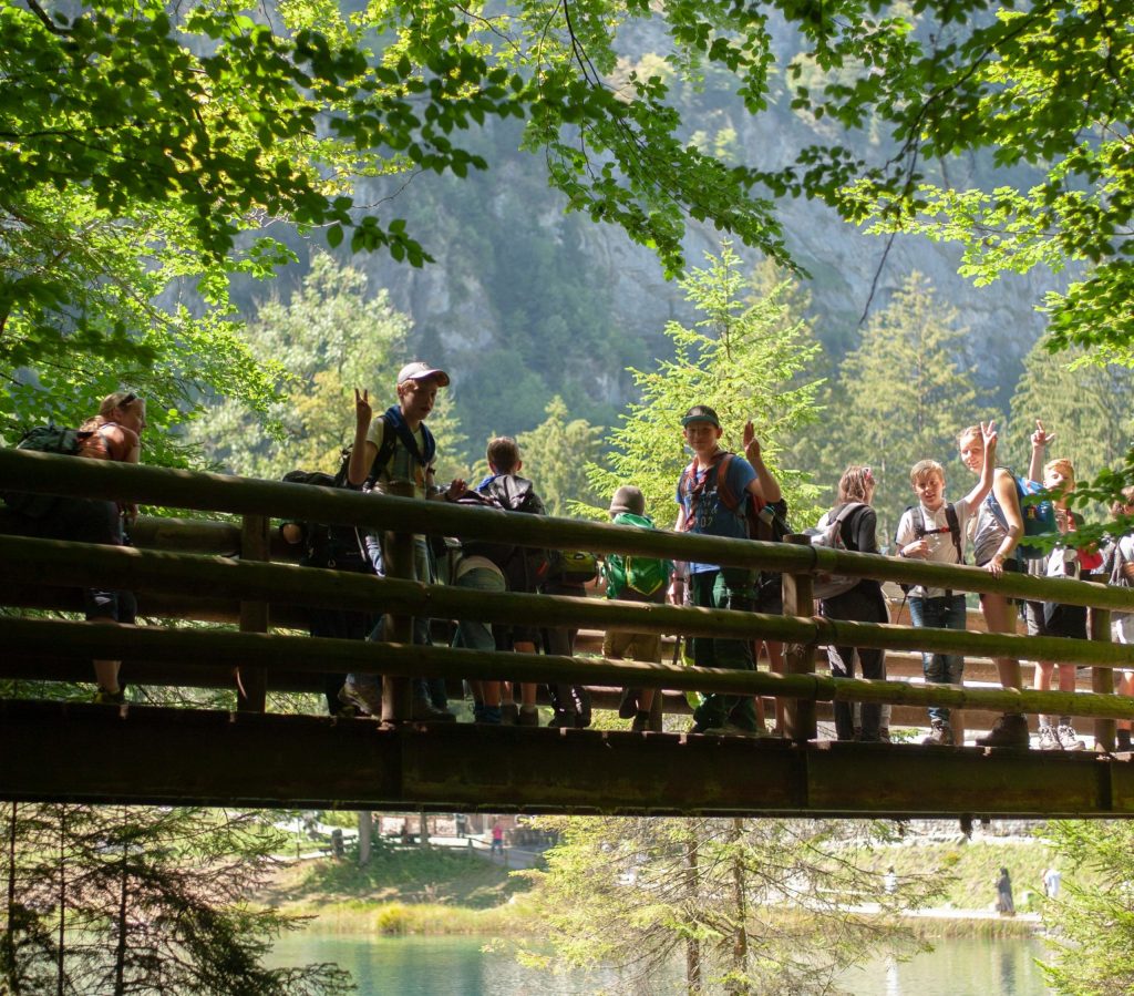 Hier stehen Pfadfinder auf einer Brücke. Der See ist grün und bewaldet.
