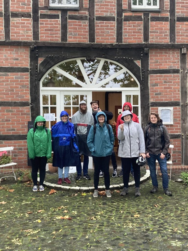 Ein Teil der LR steht in Regenjacken vor dem Eingang eines Fachwerkhauses mit roten Backsteinen.